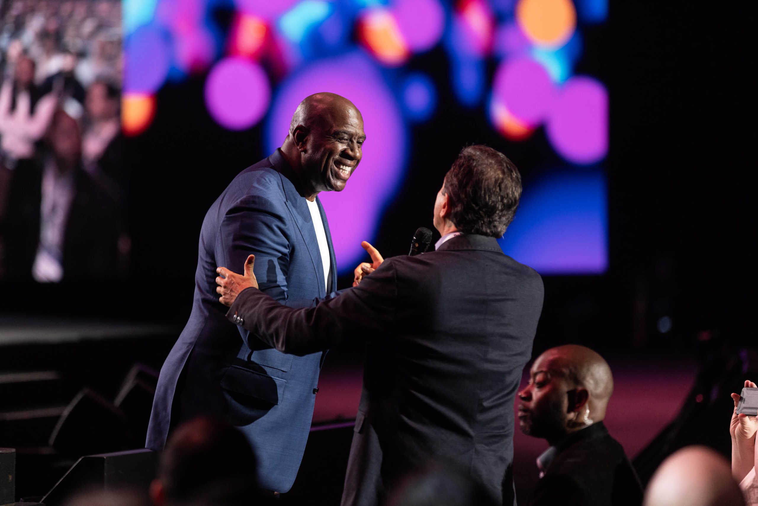 Two men in business suits greet each other with a handshake on stage an event, with colorful bokeh lights displayed on screens in the background and an audience visible in the foreground.